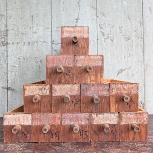 May include: A stack of small, aged wooden drawers with numbers handwritten on the front. The drawers are arranged in a pyramid shape, with a wooden knob on each drawer. The wood has a reddish-brown hue with a lighter, faded top.
