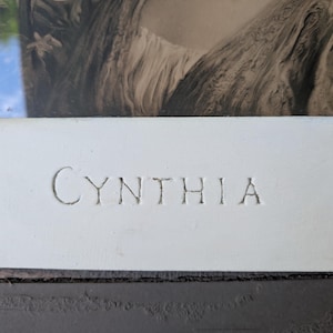 May include: A framed black and white print featuring a detailed illustration of a person with flowing hair and a draped garment. The bottom section of the frame is a wide, matte white border with the name CYNTHIA embossed in capital letters.