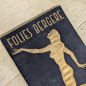 May include: A rectangular black plaque featuring the gold-colored text FOLIES BERGERE at the top. Below the text is a gold-colored embossed illustration of a figure with long hair, reaching out with one arm while looking upward.