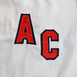 May include: Close-up of a white fabric with the letters "AC" appliqued in orange with black trim. The letters are stitched onto the fabric, creating a textured effect. The background is plain white.