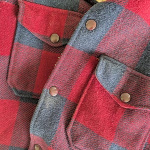 May include: A red and charcoal gray buffalo plaid shirt featuring two chest pockets with flaps, secured by dark brown metal snap buttons. The garment has a visible cream-colored fleece lining along the inner collar area, providing a textured contrast.