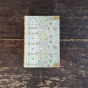 May include: A vintage book titled "Poems by Tennyson" with a floral pattern cover featuring purple, yellow, and white flowers on a light green background. The spine has a decorative design with gold accents. The book rests on a dark wooden surface.