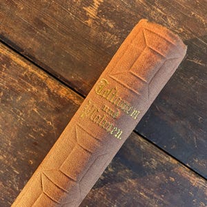 May include: A rolled-up, brown fabric item with embossed geometric patterns and gold lettering. The text reads "Gedanken und Wahrheiten." The item is resting on a dark wooden surface.