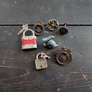 May include: An assortment of vintage hardware and decorative items, including two padlocks with keys, several drawer pulls, and a small blue rabbit figurine. The items are arranged on a dark, weathered wooden surface.