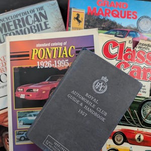 May include: A collection of vintage automotive books, including "The Encyclopedia of the American Automobile," "Standard Catalog of Pontiac 1926-1995," and "Royal Automobile Club Guide & Handbook 1957." The books feature classic car illustrations and titles.