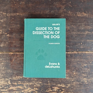 May include: A teal-colored book titled "Miller's Guide to the Dissection of the Dog" with a paw print and the text "Fourth Edition." The book is on a dark wooden surface.