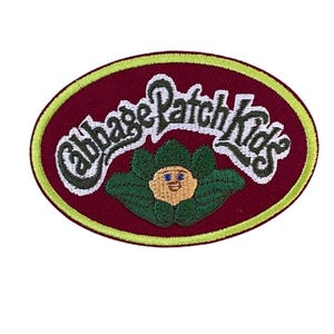 May include: An oval-shaped embroidered patch featuring a deep maroon background with a bright lime green border. The text Cabbage Patch Kids is stitched in white with dark outlines above a small character face surrounded by green leaves.