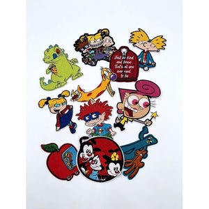 May include: A collection of embroidered patches featuring various cartoon characters including a green reptile, a yellow dog, a red-haired child, and a maroon sweater with the text Just be kind and brave That's all you ever need to be.