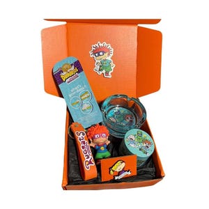 May include: An open orange gift box containing Rugrats themed items: a Dunkaroos snack package, a clear glass ashtray, a small round container, a Rugrats keychain, a character sticker, and a rectangular card featuring a cartoon character illustration.