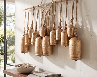 Puede incluir: Una decoración de pared que presenta doce campanas de metal dorado desgastado de varios tamaños, suspendidas por gruesas cuerdas de yute de una barra de madera horizontal, con un elemento central de macramé color crema.