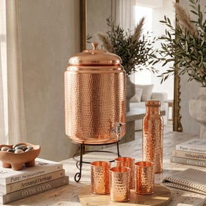 May include: A large hammered copper water dispenser with a lid and spigot on a black metal stand, accompanied by a matching hammered copper bottle and four small hammered copper tumblers arranged on a round wooden board for serving.