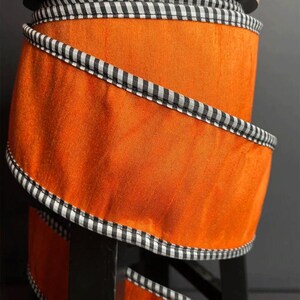 May include: A spool of orange ribbon with a black and white checkered trim. The ribbon is wrapped around a cardboard core and is displayed on a black stand. The ribbon's texture is smooth and shiny.
