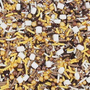 May include: A close-up view of a mix of colorful sprinkles. The mix includes white, yellow, brown, and gold sprinkles in various shapes, including rods, balls, and small squares. The sprinkles are scattered, creating a textured and visually appealing pattern.