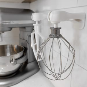 Puede incluir: Una batidora de pie plateada con un tazón de acero inoxidable y un gancho para masa blanco, junto a dos ganchos de pared blancos que sostienen un batidor plano blanco y un batidor de varillas de acero inoxidable para la batidora.
