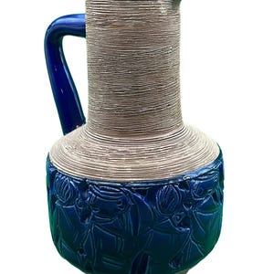 Puede incluir: Una jarra de cerámica con asa y base azules, y cuerpo beige texturizado. La base presenta un diseño en relieve detallado en azul. La jarra tiene un pico vertedor.