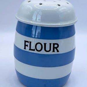 Puede incluir: Un salero de harina de cerámica con tapa blanca y cuerpo a rayas azules y blancas. La palabra "FLOUR" está impresa en negro en la parte delantera. El salero tiene un diseño clásico y vintage.