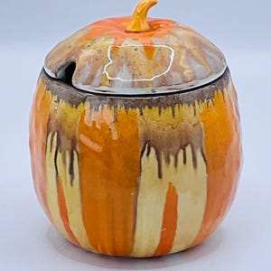 Puede incluir: Un tarro de cerámica con forma de calabaza con tapa y asa pequeña. El tarro está pintado con rayas verticales y goteos naranjas, amarillas y marrones. La tapa es naranja y marrón con un asa pequeña y curvada.