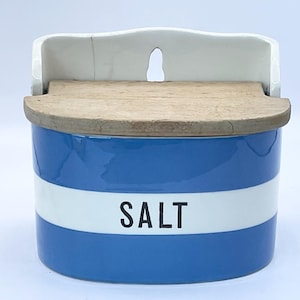 Puede incluir: Un salero de cerámica azul y blanco con tapa de madera. La palabra "SALT" está impresa en negro sobre una banda blanca. El salero tiene una parte trasera de cerámica blanca con una abertura en forma de ojo de cerradura.