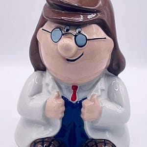 Puede incluir: Figura de cerámica de un científico de dibujos animados con cabello castaño, gafas y corbata roja. El científico lleva una bata de laboratorio blanca y un mono azul, con los pulgares hacia arriba. La figura tiene un acabado brillante y está sentada.