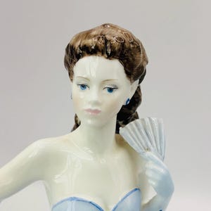May include: A porcelain figurine of a woman in a light blue dress, holding a fan. The figurine has brown hair, blue eyes, and blue earrings. The dress has a sweetheart neckline.