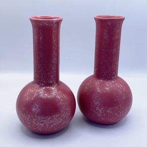 May include: Two matching, maroon-colored vases with a speckled, textured finish. Each vase has a rounded base, a long, cylindrical neck, and a flared opening. The vases are displayed against a white background.