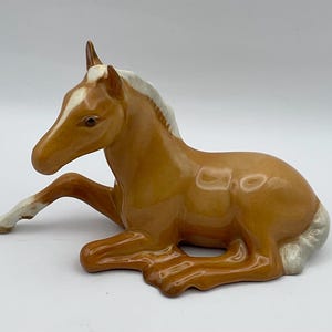 May include: A ceramic figurine of a resting foal. The horse is a warm brown color with a white blaze on its face and white on its mane and legs. The figurine is lying down with its legs tucked beneath it.