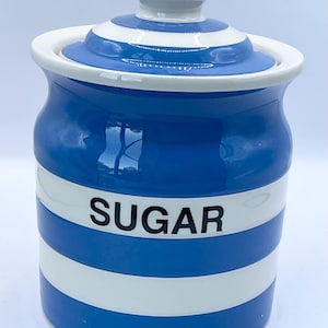 Peut inclure: Un pot à sucre en céramique à rayures bleues et blanches avec un couvercle. Le pot a une forme cylindrique avec une bande blanche au milieu et le mot "SUGAR" en noir. Le couvercle est également à rayures bleues et blanches avec un petit bouton blanc.