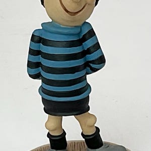 Könnte beinhalten: Figur einer lächelnden Cartoonfigur mit schwarzen Haaren und einer roten Schleife. Die Figur trägt ein blau-schwarz gestreiftes Hemd, schwarze Shorts und graue Schuhe. Die Figur steht auf einem runden, cremefarbenen Sockel.