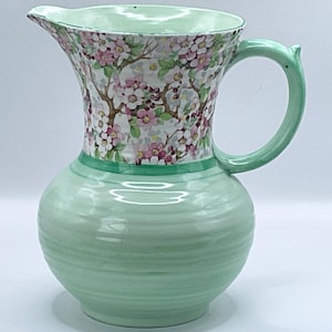 May include: A vintage ceramic jug with a light green base and handle. The upper section displays a floral pattern of pink and white blossoms on a cream backdrop. The jug has a spout and a rounded body.