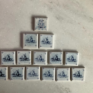 May include: A collection of square ceramic tiles with blue and white illustrations. The tiles feature images of windmills and sailing ships. The tiles are arranged in a pyramid shape on a light-coloured surface.