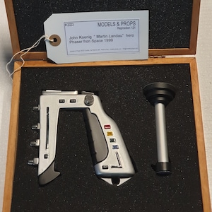Könnte beinhalten: Eine silberne und schwarze Requisiten-Phaser-Pistole und ein zylindrisches Objekt, wahrscheinlich eine Requisite, werden in einer Holzkiste präsentiert. Der Phaser hat ein markantes eckiges Design mit farbigen Knöpfen. Ein Etikett lautet "Models & Props".