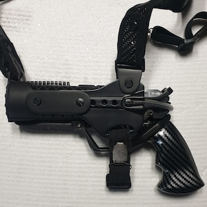 Könnte beinhalten: Ein schwarzes taktisches Schulterholster-System mit einem geformten Polymerrahmen, verstellbaren Nylongurten und einem Griff mit Kohlefaser-Muster. Das Holster verfügt über einen sicheren Haltemechanismus, eine Metall-Einstellschraube und Gurte.