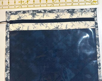 May include: A quilted project bag with a clear vinyl window. The bag features a navy blue floral print, a cream-colored floral accent, and a navy zipper. A butterfly-shaped pin rests on a yellow grid ruler.
