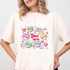 May include: Ivory-colored t-shirt featuring a colorful graphic of heart-shaped candies with positive affirmations like "Loved," "Beautiful," and "Worthy." The words "You are" are in the center. The shirt has short sleeves and a relaxed fit.
