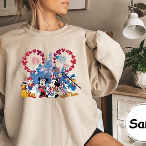 Puede incluir: Sudadera color arena con un gr&aacute;fico colorido de personajes de Disney con atuendos patri&oacute;ticos frente a un castillo y fuegos artificiales. Los personajes incluyen a Mickey Mouse, Minnie Mouse, Donald Duck y Pluto. La palabra "Sand" est&aacute; impresa abajo.