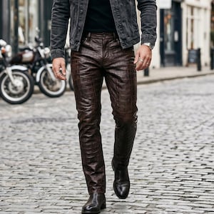 May include: Brown leather trousers with a crocodile skin texture. The trousers are worn with black boots and a dark denim jacket. The person is walking on a cobbled street with motorcycles in the background.
