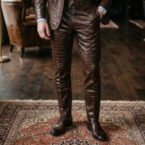 May include: A brown leather suit with a crocodile pattern. The suit comprises a blazer, waistcoat, and trousers. The person is wearing a light blue shirt and brown leather shoes. The suit is worn on a patterned rug.