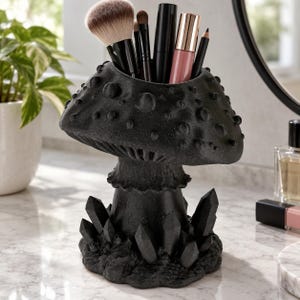 May include: A black mushroom-shaped organiser featuring a textured cap with raised spots and a base surrounded by pointed crystal-like structures. The top of the mushroom cap is open, holding various makeup brushes, a black tube, and a pink lipstick.