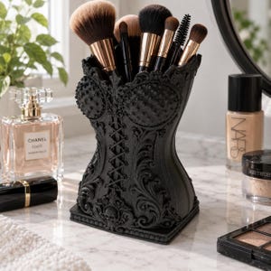 Puede incluir: Un soporte para brochas de maquillaje negro con forma de corsé, con intrincados relieves, un detalle de cordones central y un pequeño motivo de calavera en la base. El soporte contiene varias brochas con virolas de oro rosa y cerdas oscuras.