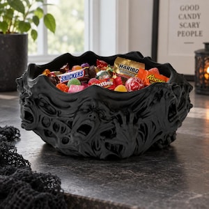 May include: A matte black bowl featuring a relief design of multiple screaming faces and reaching hands around the exterior. The bowl is filled with various colorful candy wrappers including Snickers, Haribo Goldbears, Skittles, and Reese's.