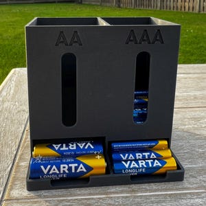 Puede incluir: Un organizador de baterías negro con compartimentos para pilas AA y AAA. El organizador tiene ranuras para facilitar el acceso y muestra varias pilas VARTA Longlife. Los textos "AA" y "AAA" son visibles.