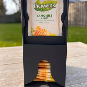 Könnte beinhalten: Ein schwarzer Teebeutelhalter aus Metall mit einem Stapel Teebeutel, der durch eine gebogene Öffnung sichtbar ist. Auf der Oberseite des Halters befindet sich ein Teebeutel "Pickwick Herbal Camomile Honey".