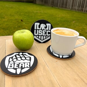 Puede incluir: Una taza de café blanca con un líquido marrón se encuentra sobre un posavasos blanco y negro. Se ven dos posavasos adicionales con texto y gráficos blancos, junto con una manzana verde. Los posavasos tienen las palabras "USEC" y "BEAR".