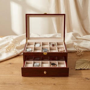 May include: A wooden watch box with a glass top and a mirrored interior. The box is divided into compartments to hold multiple watches. The drawer is open, revealing additional watch storage. The box is made of dark wood with a polished finish.