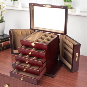 Puede incluir: Un joyero de madera teñida de color cereza oscuro con múltiples cajones y compartimentos. La caja tiene una tapa con bisagras con un espejo y compartimentos laterales para colgar collares. Los herrajes dorados acentúan los cajones y las puertas.