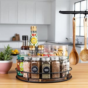 Puede incluir: Un especiero giratorio de madera y metal lleno de varios frascos, botellas y recipientes de especias. El estante incluye etiquetas para "Chili Powder" y "Celery Flakes". Cucharas de madera cuelgan en el fondo, y una pequeña planta en maceta está en la encimera.