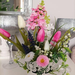 Może przedstawiać: Kompozycja kwiatowa z różowymi tulipanami, hiacyntami i stokrotkami w białym doniczce z koronkową krawędzią. Zielone liście i gałązki białych kwiatów dopełniają aranżację. Kompozycja ustawiona jest na odblaskowym srebrnym tle.