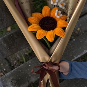 Könnte beinhalten: Ein Blumenstrauß aus handgefertigten Pfeifenreinigern mit einer großen orangefarbenen Sonnenblume mit dunkelbrauner Mitte, kleinen weißen Gänseblümchen und grünen Spiralstielen. Das Arrangement ist in bedrucktes beiges Papier gewickelt und mit einem braunen Band gebunden.