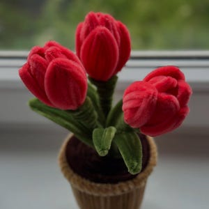 May include: A decorative arrangement of three red tulip flowers in a brown pot. The tulips have a velvety texture and green stems and leaves. The pot is a light brown colour with a woven design.