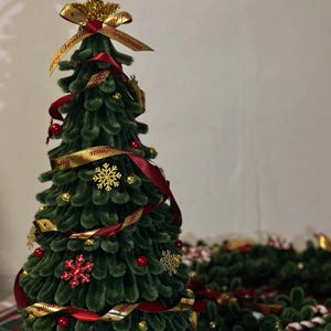 Puede incluir: Un árbol de Navidad decorativo con una superficie verde texturizada, adornado con cintas rojas, adornos dorados y detalles de copos de nieve. Una estrella dorada y una cinta con el texto "Merry Christmas" coronan el árbol. Está sobre una base blanca y marrón.